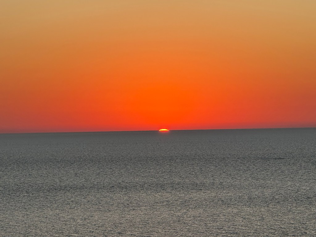 A serene sunset over the ocean, with the sun setting on the horizon and casting a warm orange glow on the water.